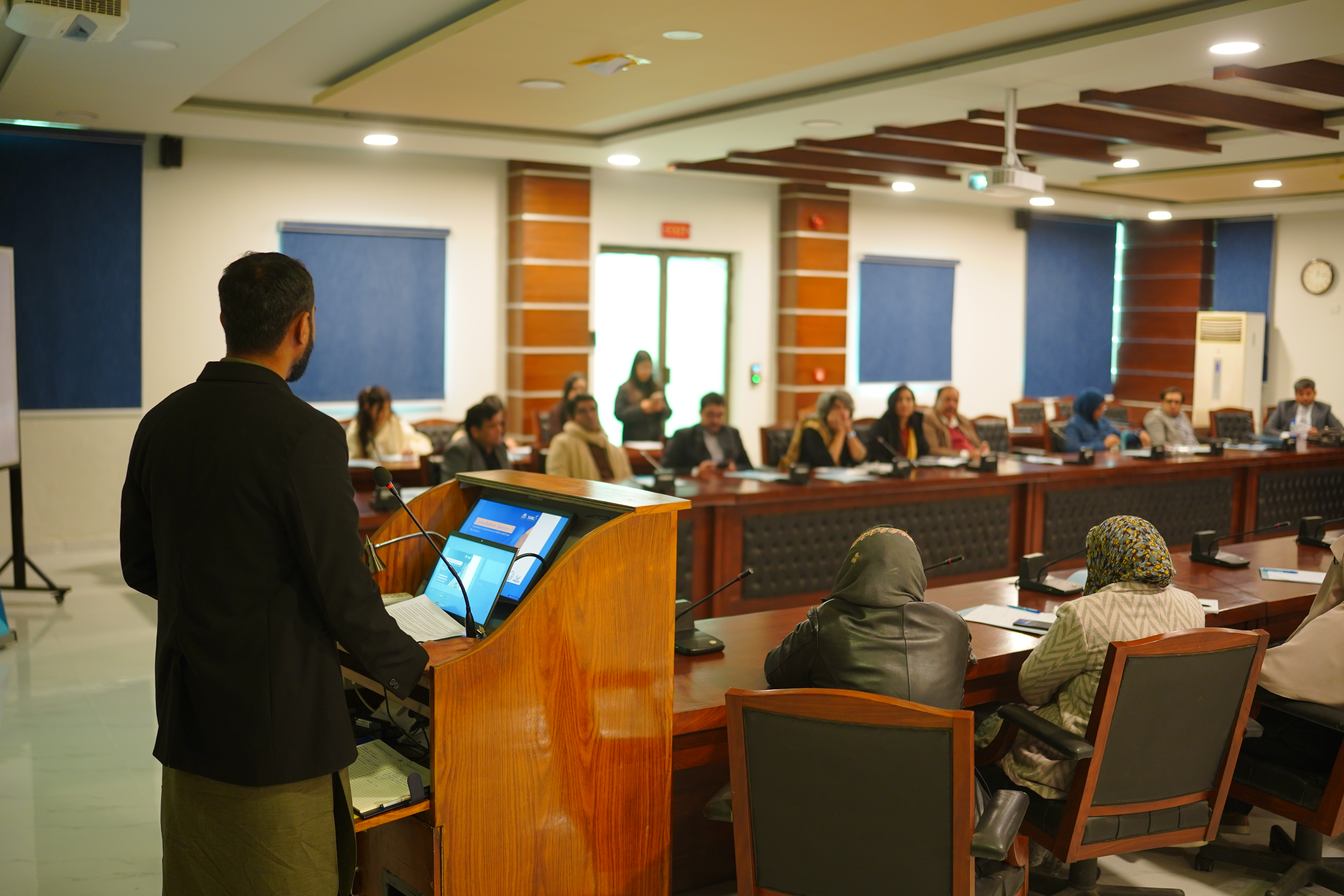A photograph of a man from behind giving a lecture to a diverse group of people