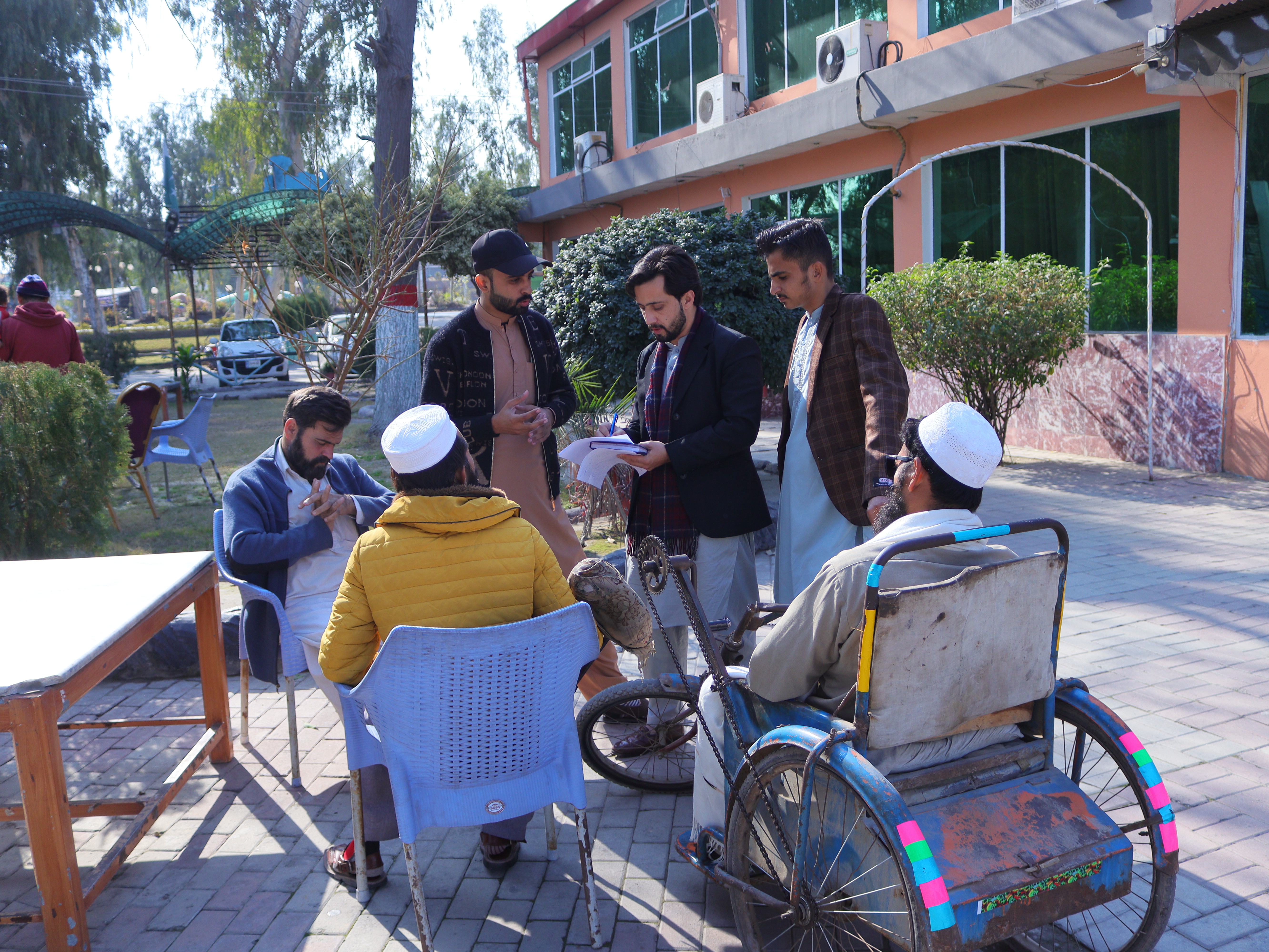 6 men, 2 seated and 1 using a wheelchair, in a circle discussing a document one of the standing men is reading from
